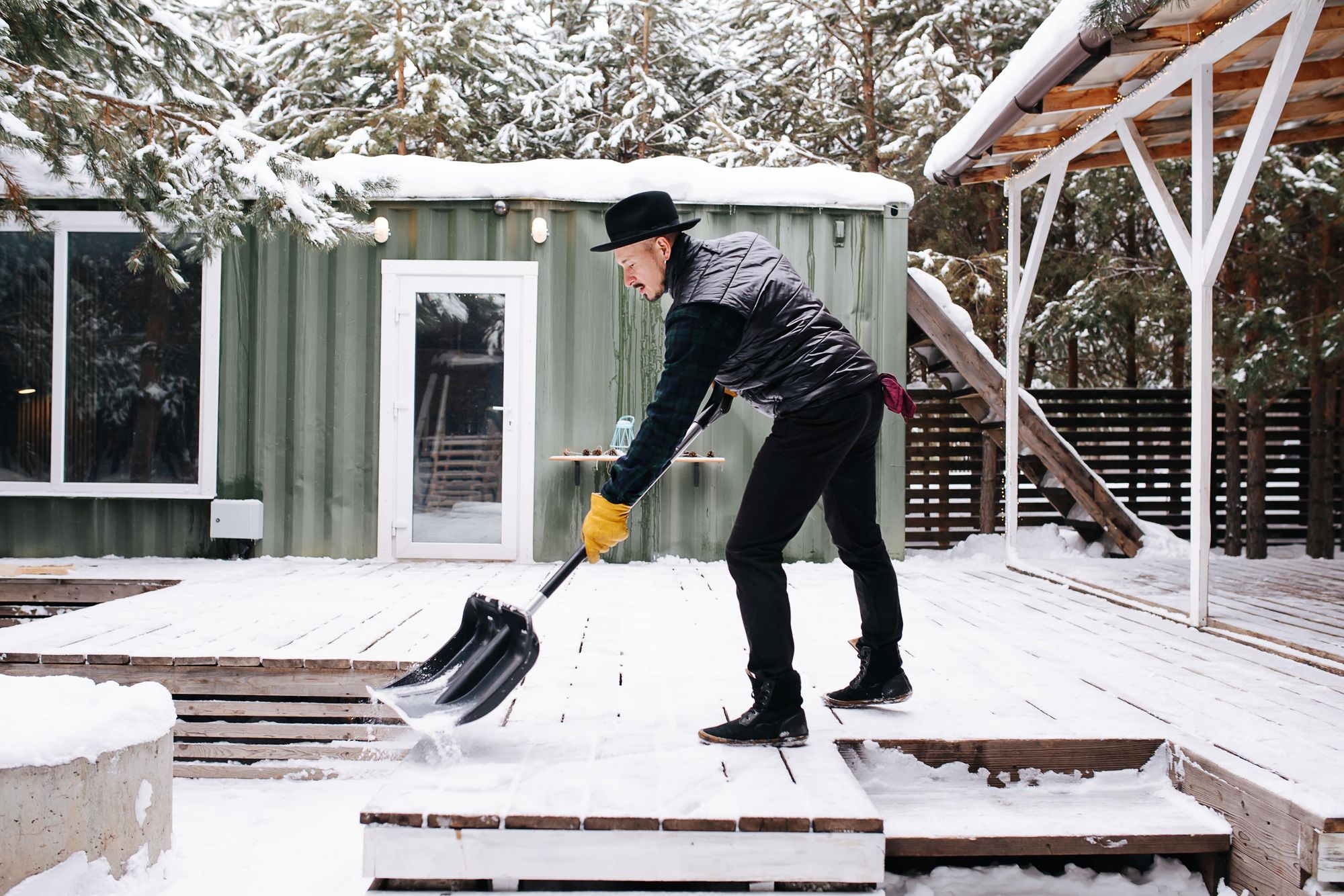 man-hat-leather-jacket-clearing-snowy-plank-floor-front-house-with-plastic-wide-shovel-min